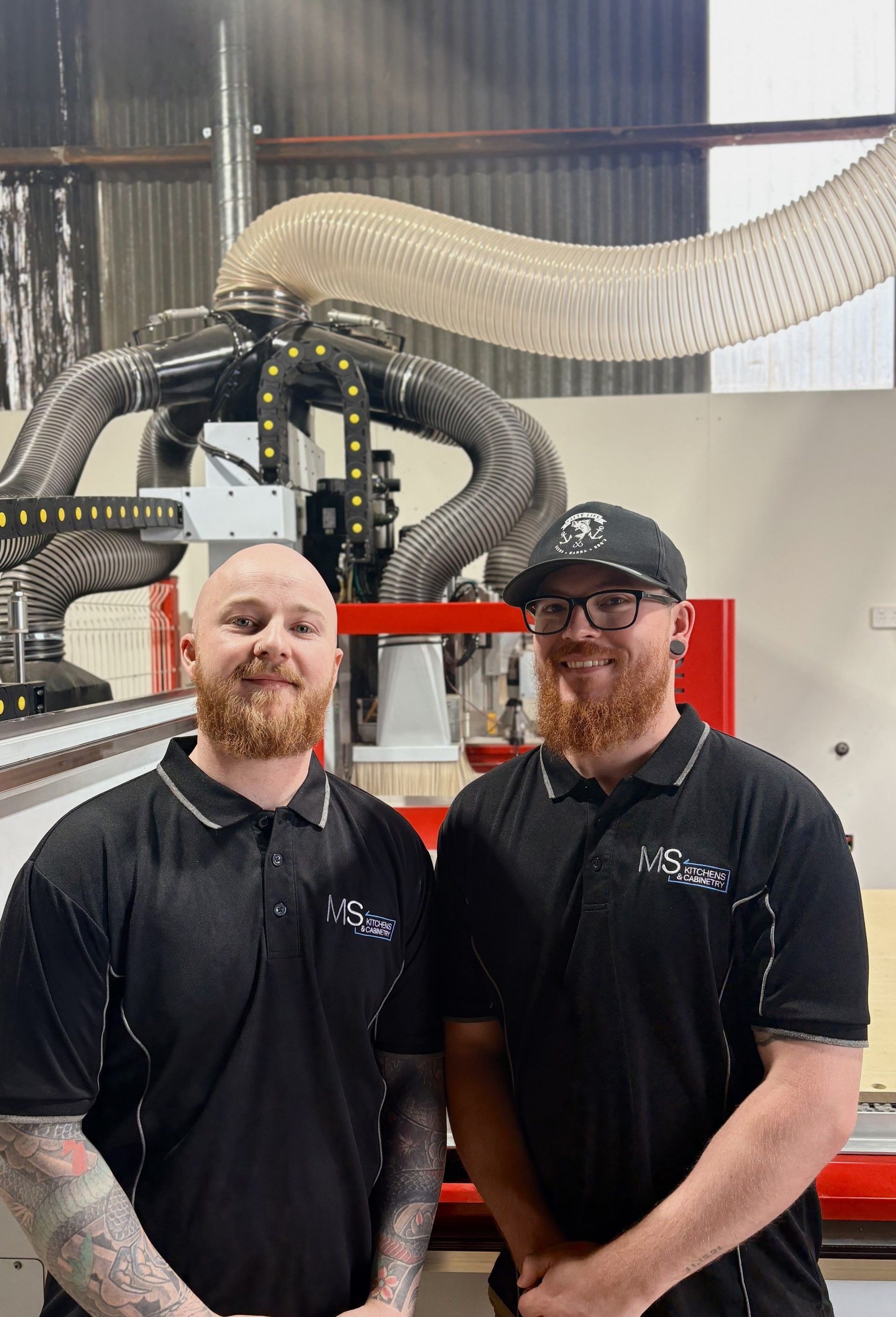 Two men with red beards, in black shirts, standing in a workshop. A large machine is behind them.