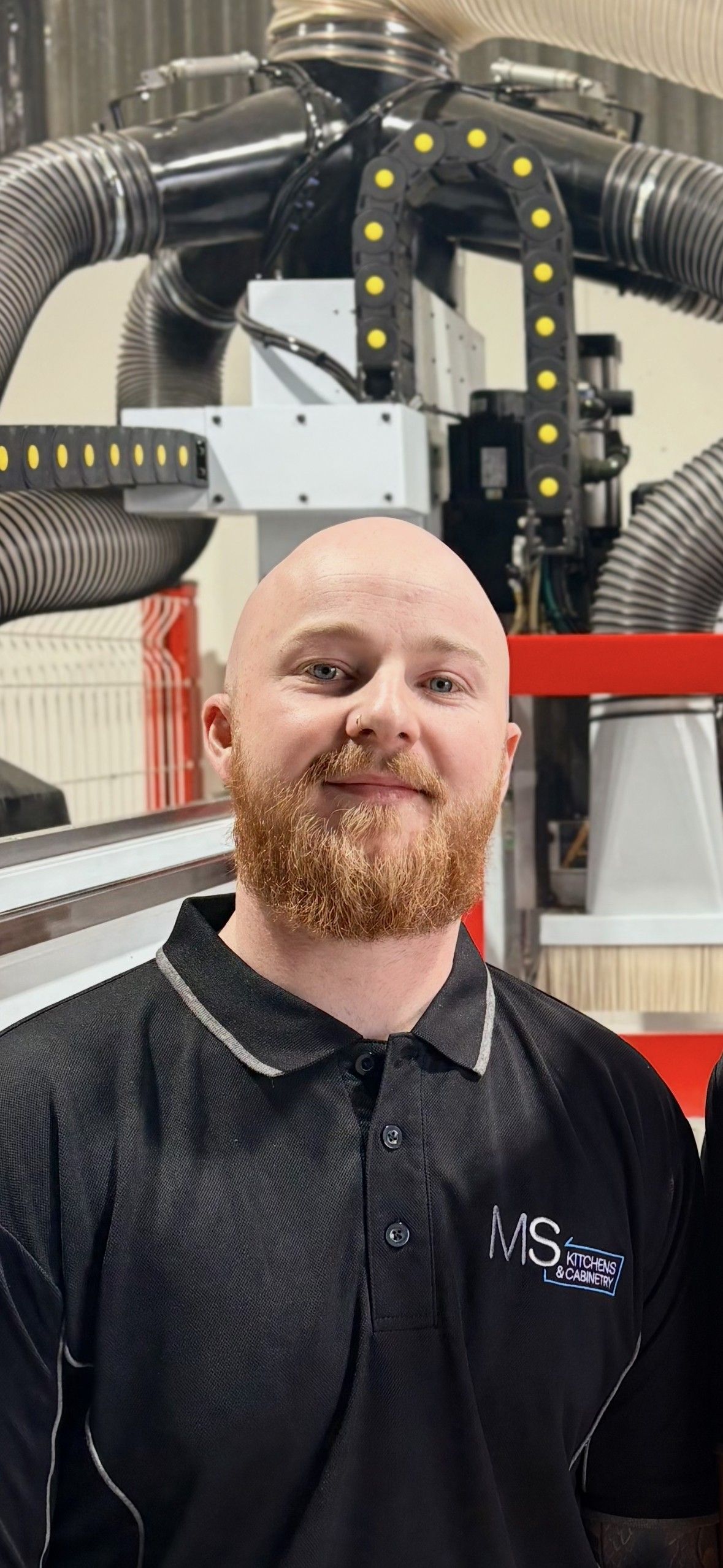 Bald man with red beard smiles, wearing a black collared shirt in a workshop with machinery.