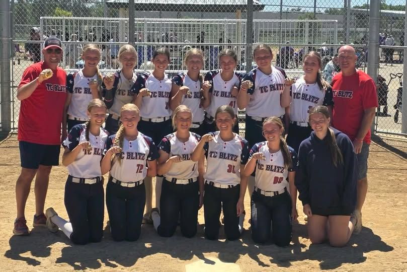 Softball team posing with coaches on a dirt field in front of a chain-link fence