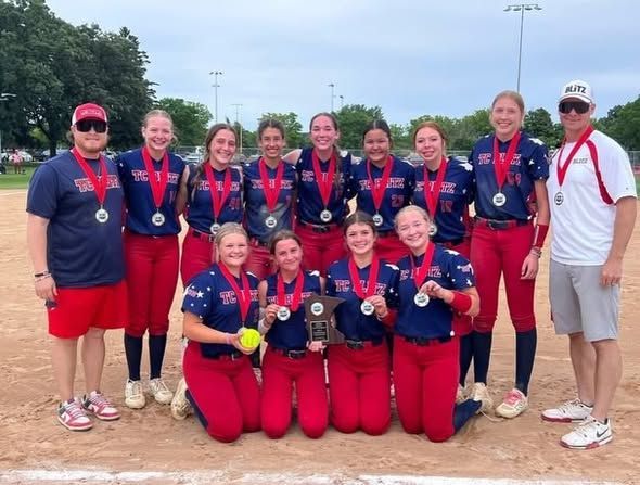 Softball team posing on a sunny field in navy and red uniforms, wearing sunglasses and smiling.