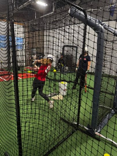 Indoor batting cage with a person in a red shirt swinging a bat on turf.