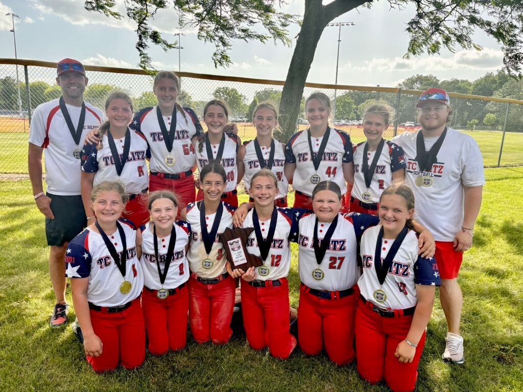 Team posing outdoors on grass, wearing medals and red-white uniforms, holding a trophy.