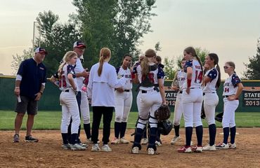 Softball team huddles with coach on dirt field at sunset, wearing white uniforms and catching gear