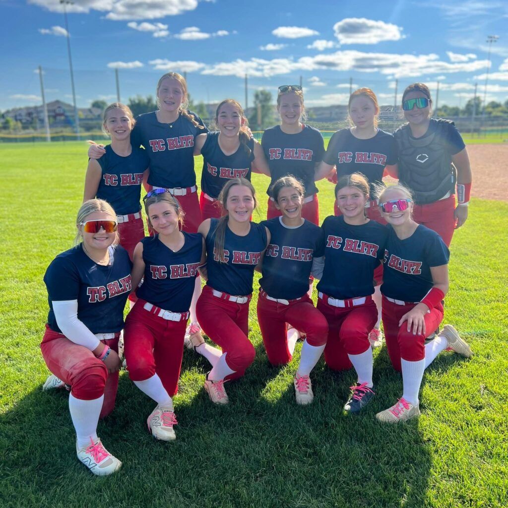 Softball team posing on a sunny field in navy and red uniforms, wearing sunglasses and smiling.