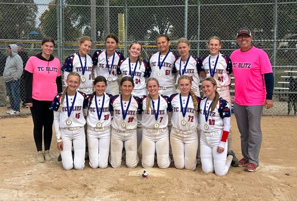 Youth softball team posing with medals on a dirt field, coaches standing behind them.