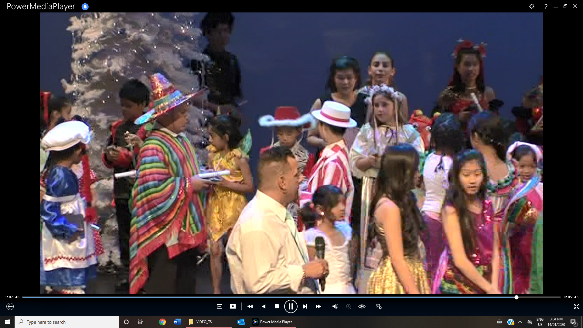 A group of children in colorful costumes are on stage with adults at a performance. Stage is decorated with a tree.