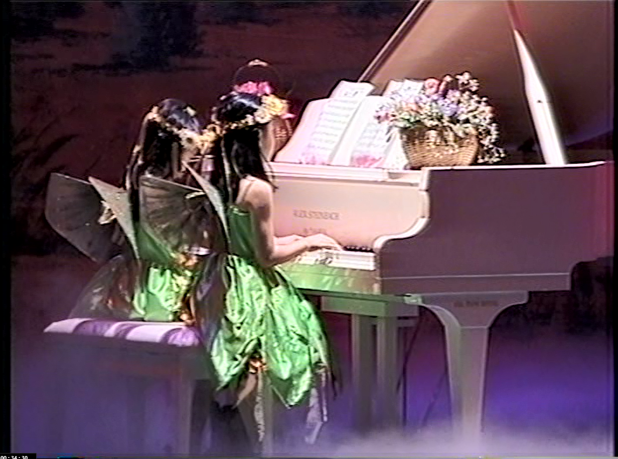 Two girls in fairy costumes play a white piano. They wear green dresses, wings, and flower crowns, with a floral arrangement on the piano.