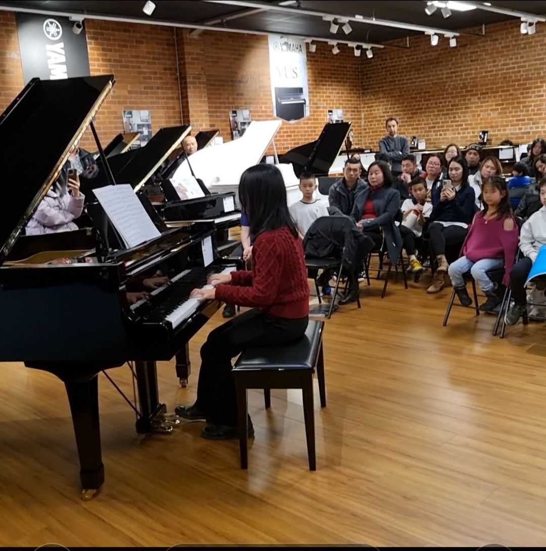 A woman plays piano in front of an audience in a music store. People watch and some clap, while others are seated at additional pianos.