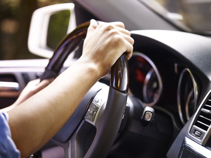 Mani che stringono il volante all'interno di un'auto, con cruscotto e specchietto laterale visibili.