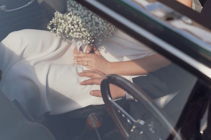 Una persona tiene in mano un piccolo mazzo di fiori bianchi all'interno di un'auto, vista attraverso il parabrezza.