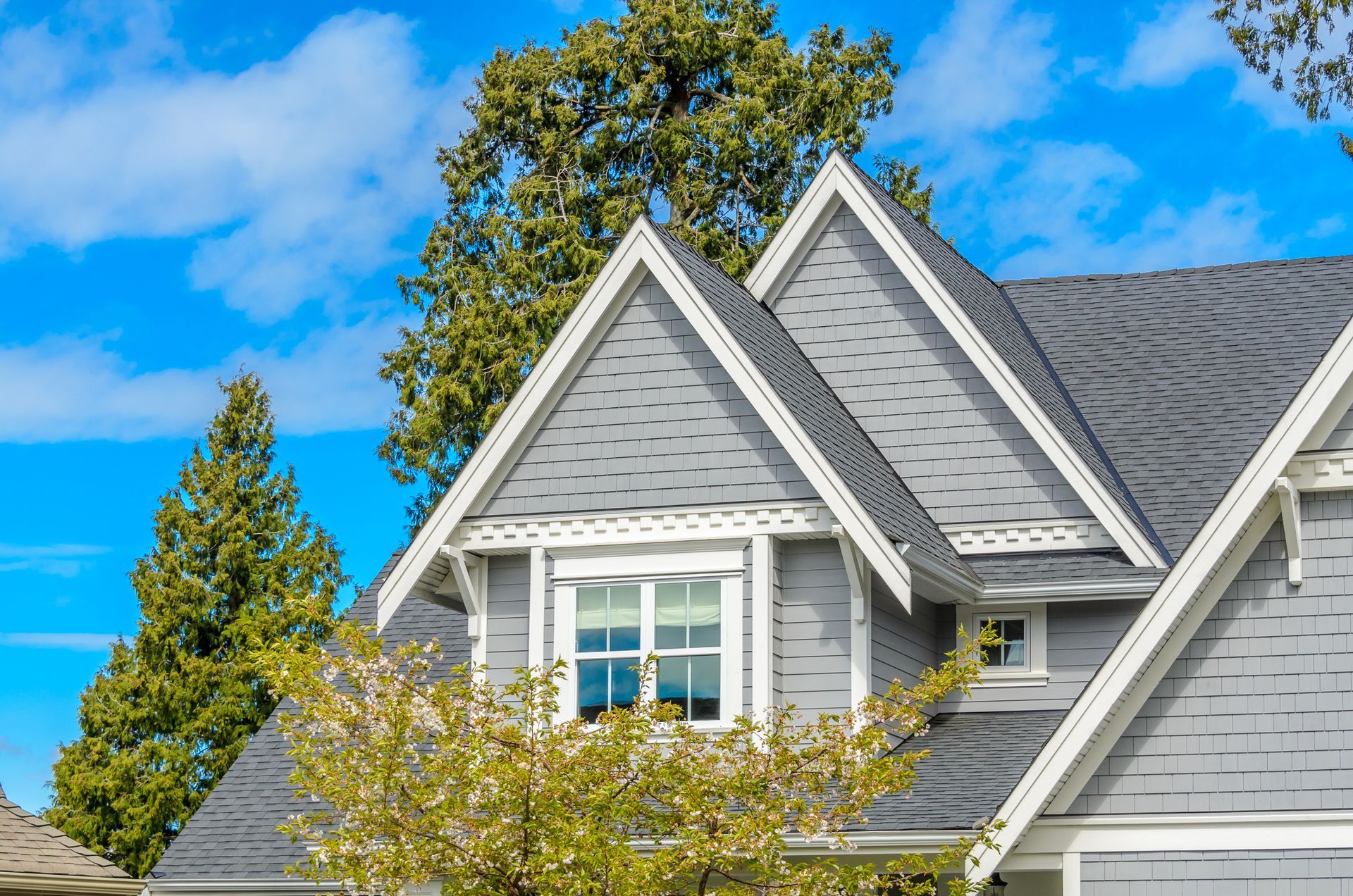 Gray house with white trim, dormer with window, blue sky, and green trees.