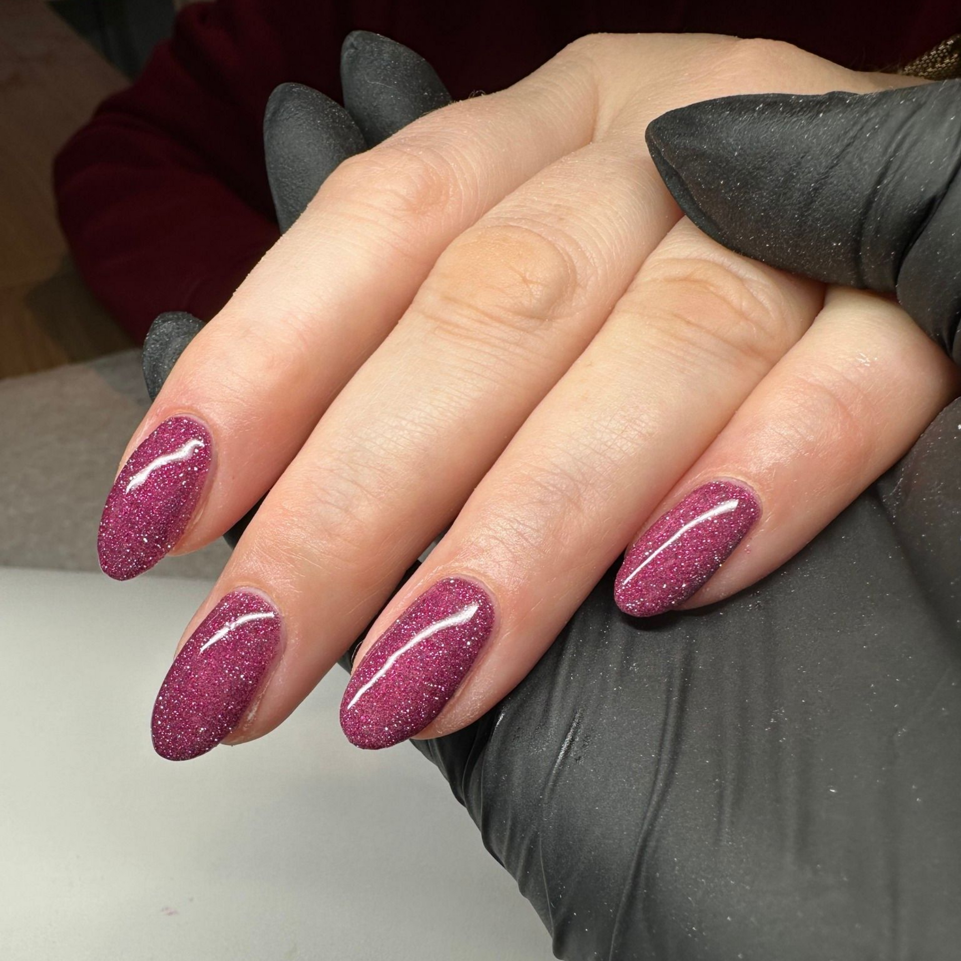 Close-up of a hand with oval-shaped nails painted a sparkly magenta color. The hand is resting in a black glove.