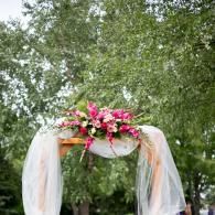 Flower-decorated wedding arch with white drapery and pink bouquet outdoors under trees