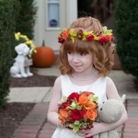 Child in white dress holding flowers and a pumpkin at a front door, with fall decorations in the background
