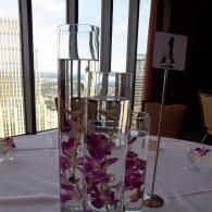 Tall glass flower vases with purple orchids on a white table by large windows with a city view