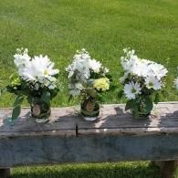 Three white flower bouquets in glass jars on a wooden bench beside green grass