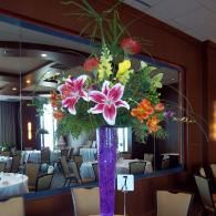 Tall purple floral arrangement with pink lilies and orange flowers in a restaurant lobby