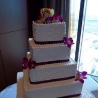 Three-tier white wedding cake with purple flowers, displayed by a window