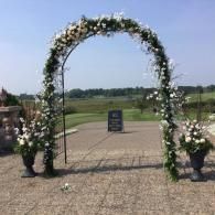 Floral archway decorated with white flowers and greenery on a paved outdoor path overlooking fields