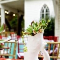 White pitcher with green succulents on a colorful café table, blurred outdoor seating in the background