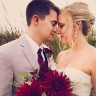 Bride and groom touching foreheads, holding a deep red bouquet outdoors.