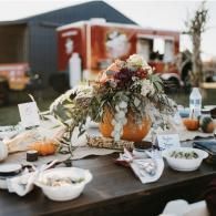 Outdoor harvest-style table with pumpkins, flowers, and dishes set in front of a barn