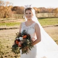 Bride in a white lace gown holding a bouquet outdoors in a field, smiling in the sunlight