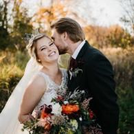 Bride and groom smiling outdoors with a bouquet, the groom kissing her cheek