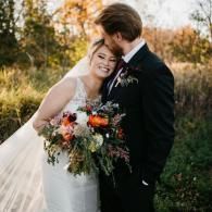 Bride and groom embrace outdoors, smiling with a colorful bouquet in autumn light