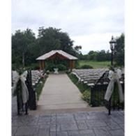 Outdoor wedding aisle with white chairs and floral decorations leading to a small gazebo, framed by greenery