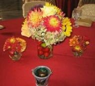 Three small flower arrangements on a red tablecloth, with a larger red-and-yellow bouquet in the center.