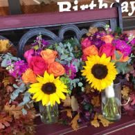 Two bright floral bouquets with sunflowers and roses in glass vases on a table under a birthday sign