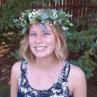 Smiling person wearing a white floral crown and black patterned top outdoors in front of greenery