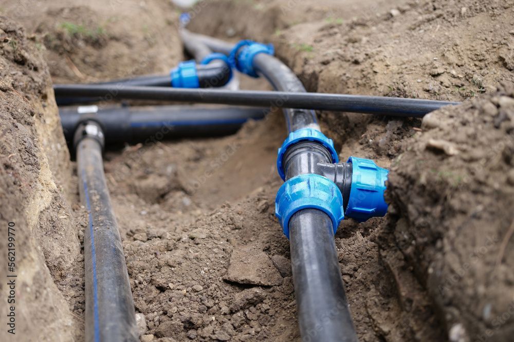 Man working on irrigation system valves in a buried box surrounded by soil and black tubing. Black pipes with blue connectors installed in a trench, likely for a water system.