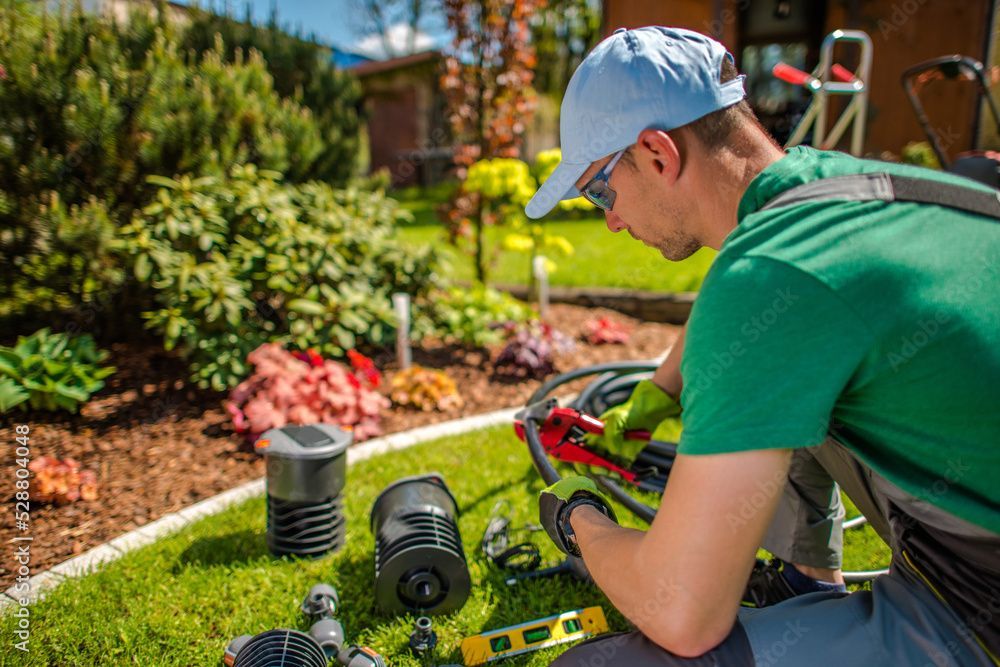Gardener in green shirt and blue cap working on irrigation system parts in a landscaped yard.