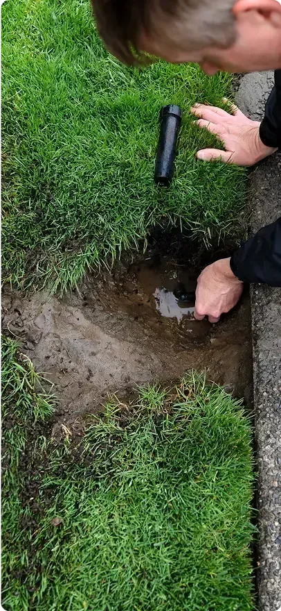 Person examines a sprinkler head in a patch of dirt surrounded by green grass.