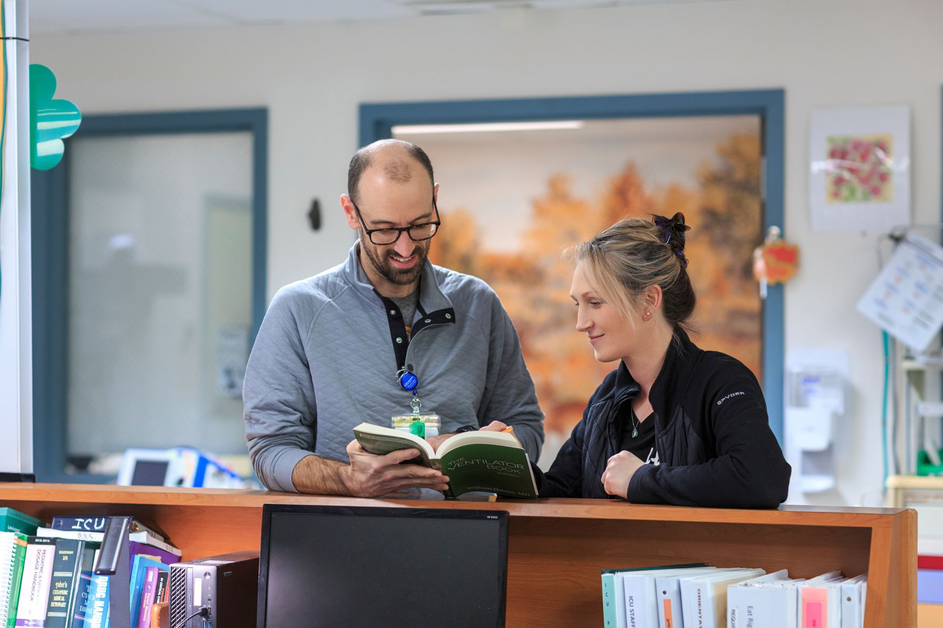 Two health professionals, a man and a woman, reading a book together at a hospital reception desk.