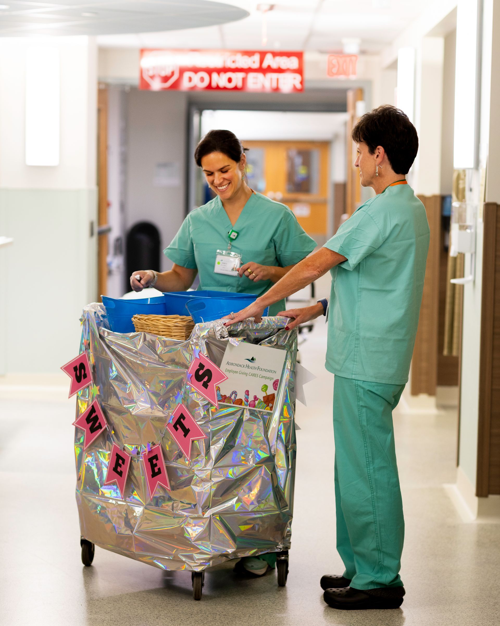 Two nurses in scrubs decorate a supply cart with sweets in a hospital hallway.