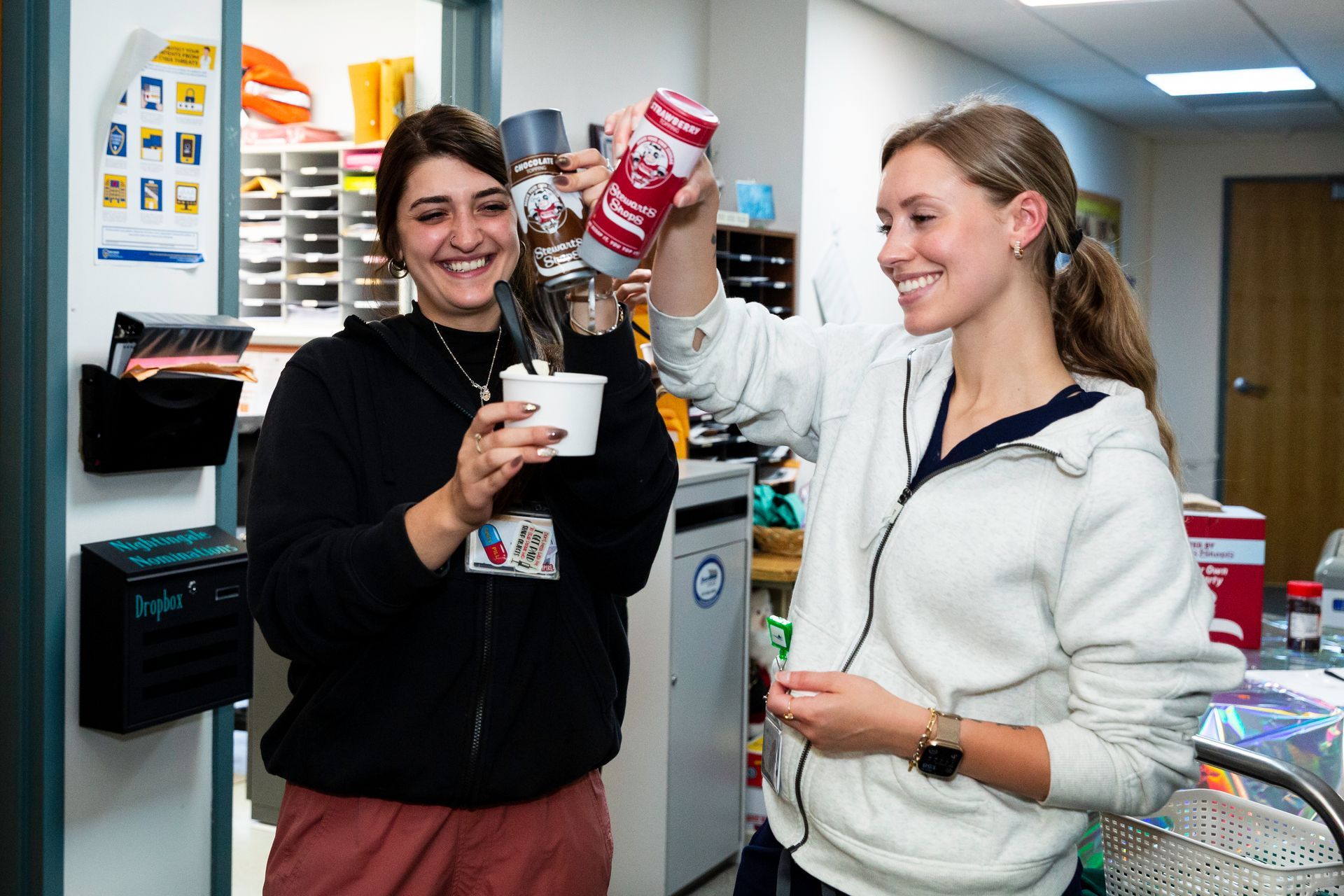 Two women smiling, one adding whipped cream to a cup, in a brightly lit office.