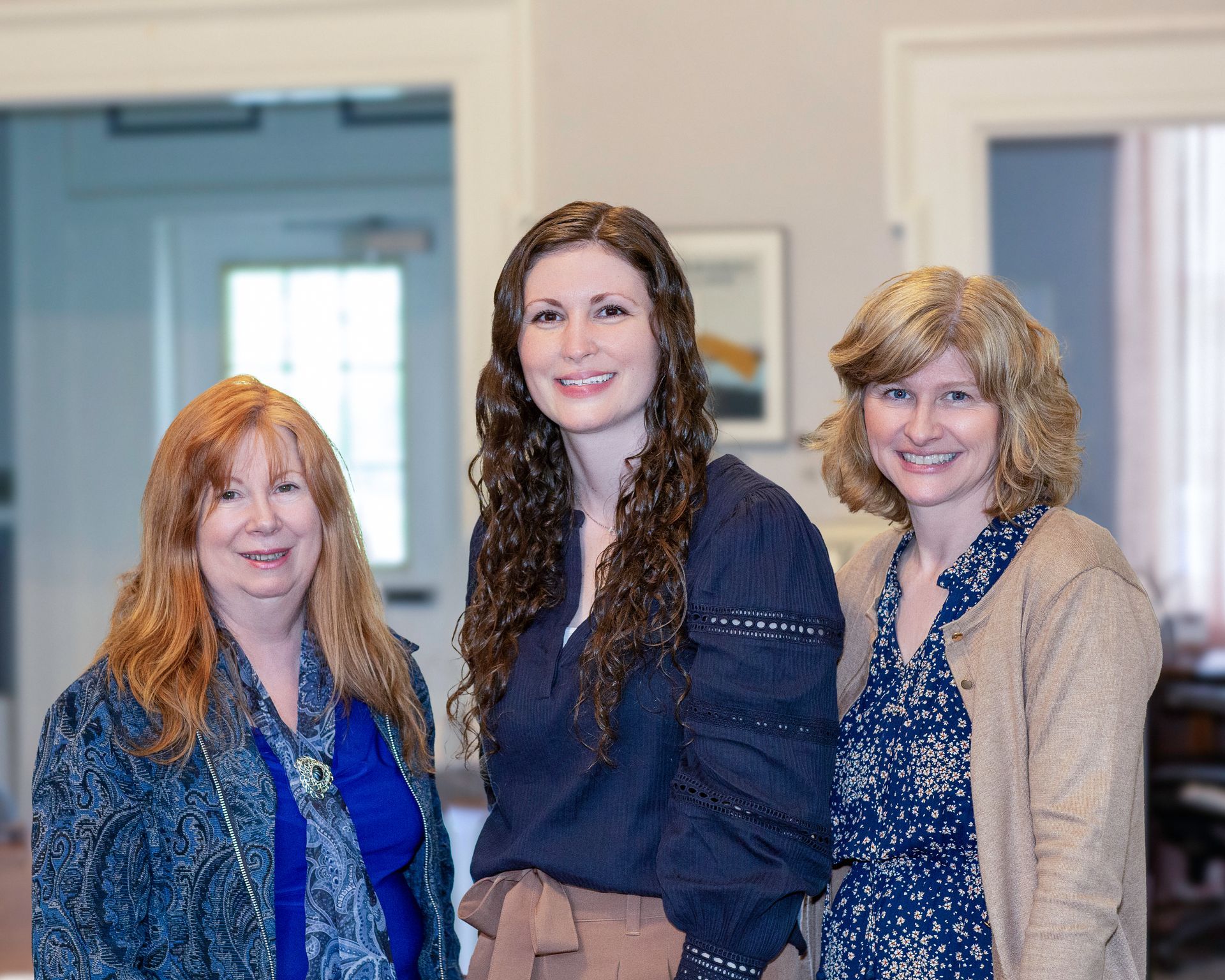 Three women smiling indoors: One with red hair, one with long brown hair, one with blonde hair.