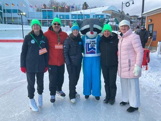 Group of six people, including a raccoon mascot, on an ice skating rink, all wearing green hats.
