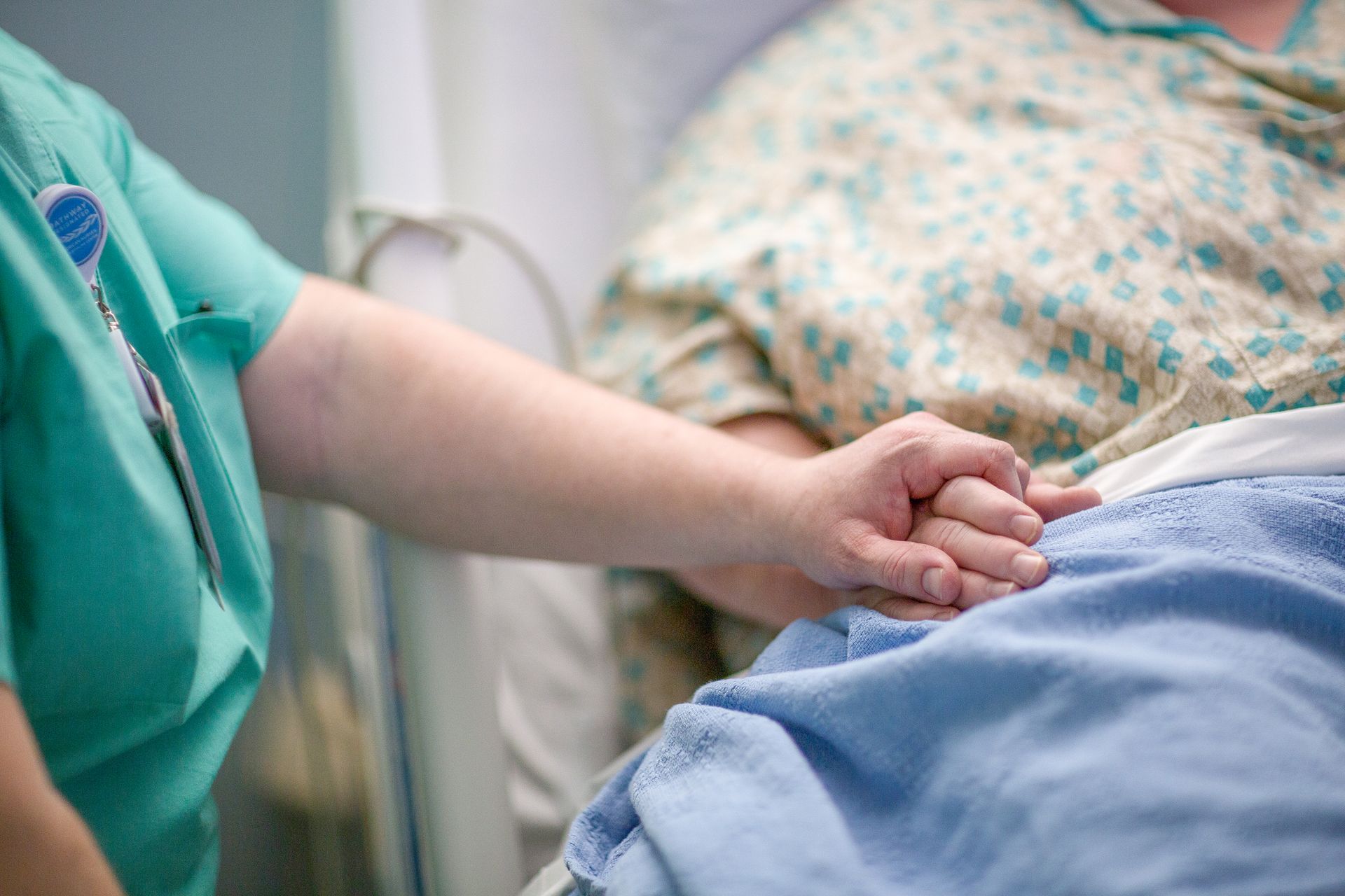 Nurse in green scrubs holds the hand of a patient in a hospital bed, offering comfort.
