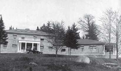 Black and white photo of a large brick house with a porch, a parked car, and trees in the background.