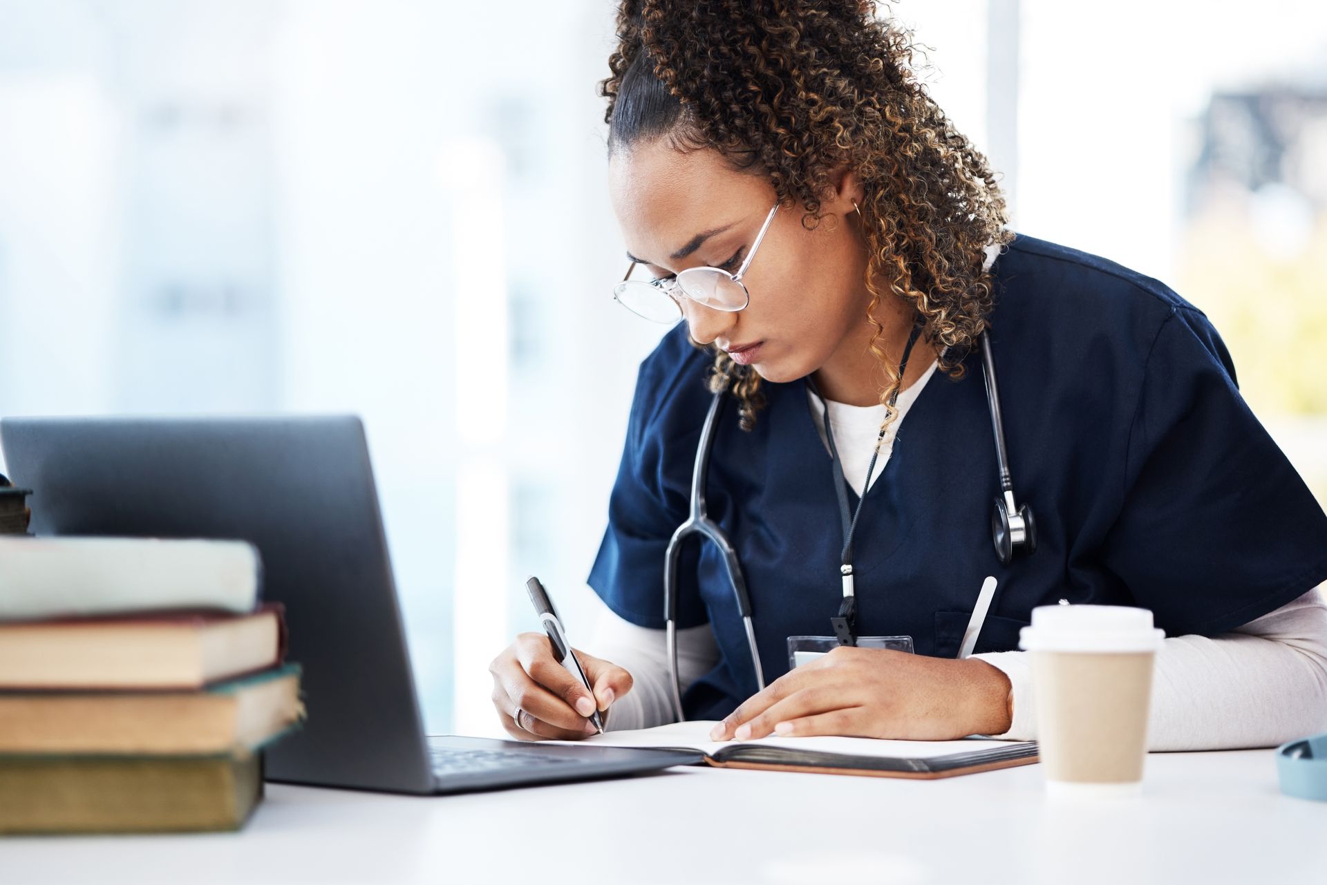 A medical professional with glasses, writing in a notebook, studying, laptop and coffee on the desk.