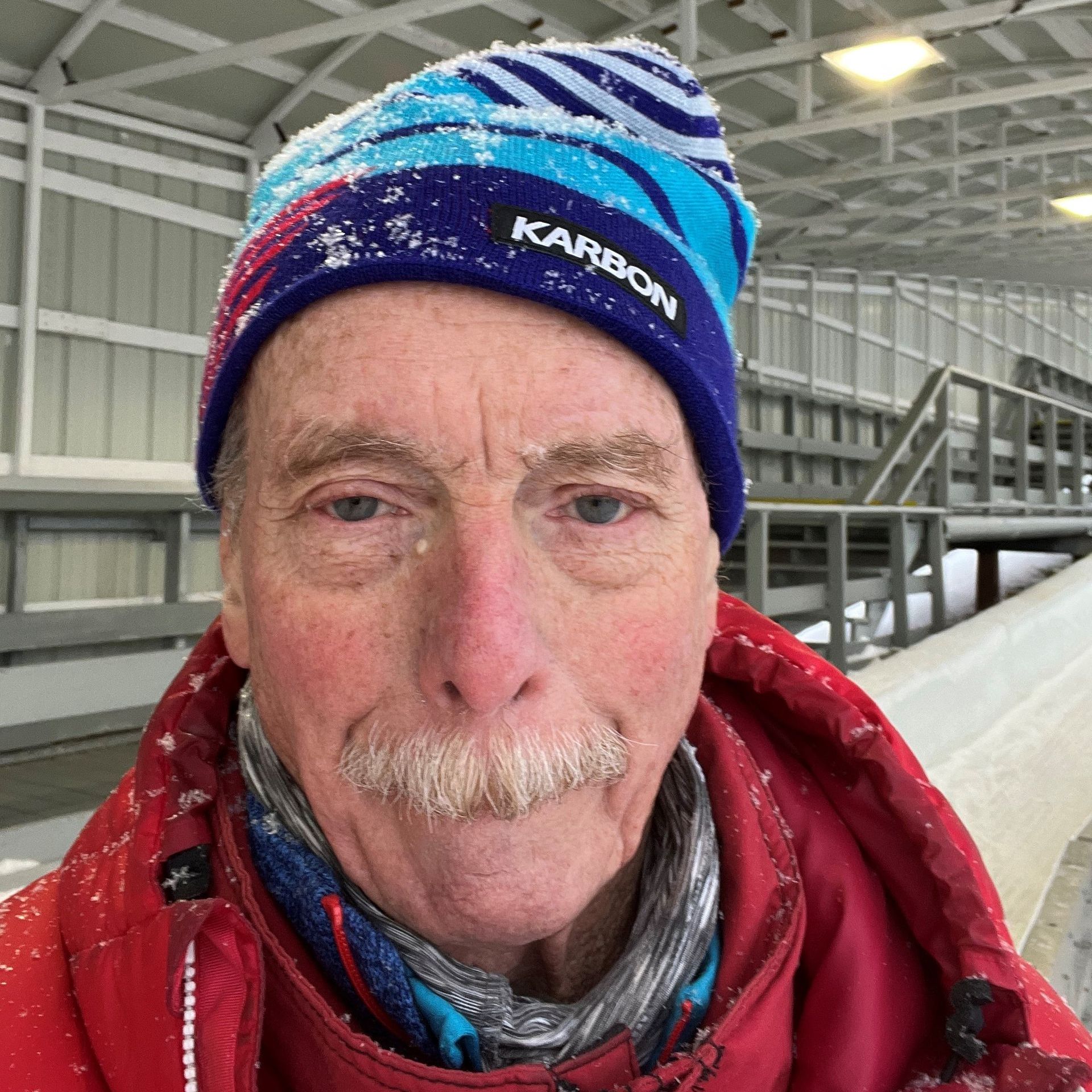 Elderly man in a blue and red winter hat and coat, smiling slightly, dusted with snow.