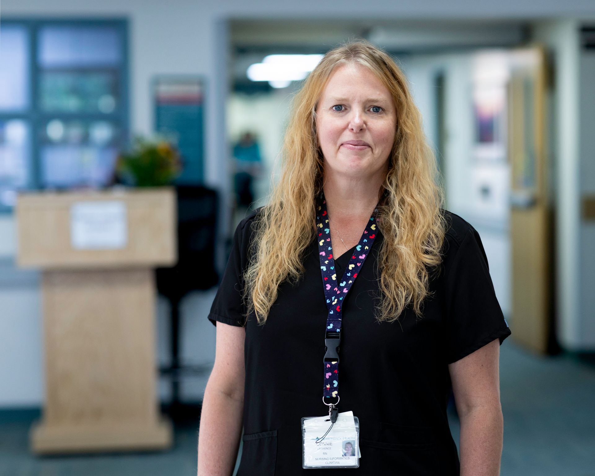 Woman with blonde hair wearing a black shirt and lanyard stands in a hospital hallway.