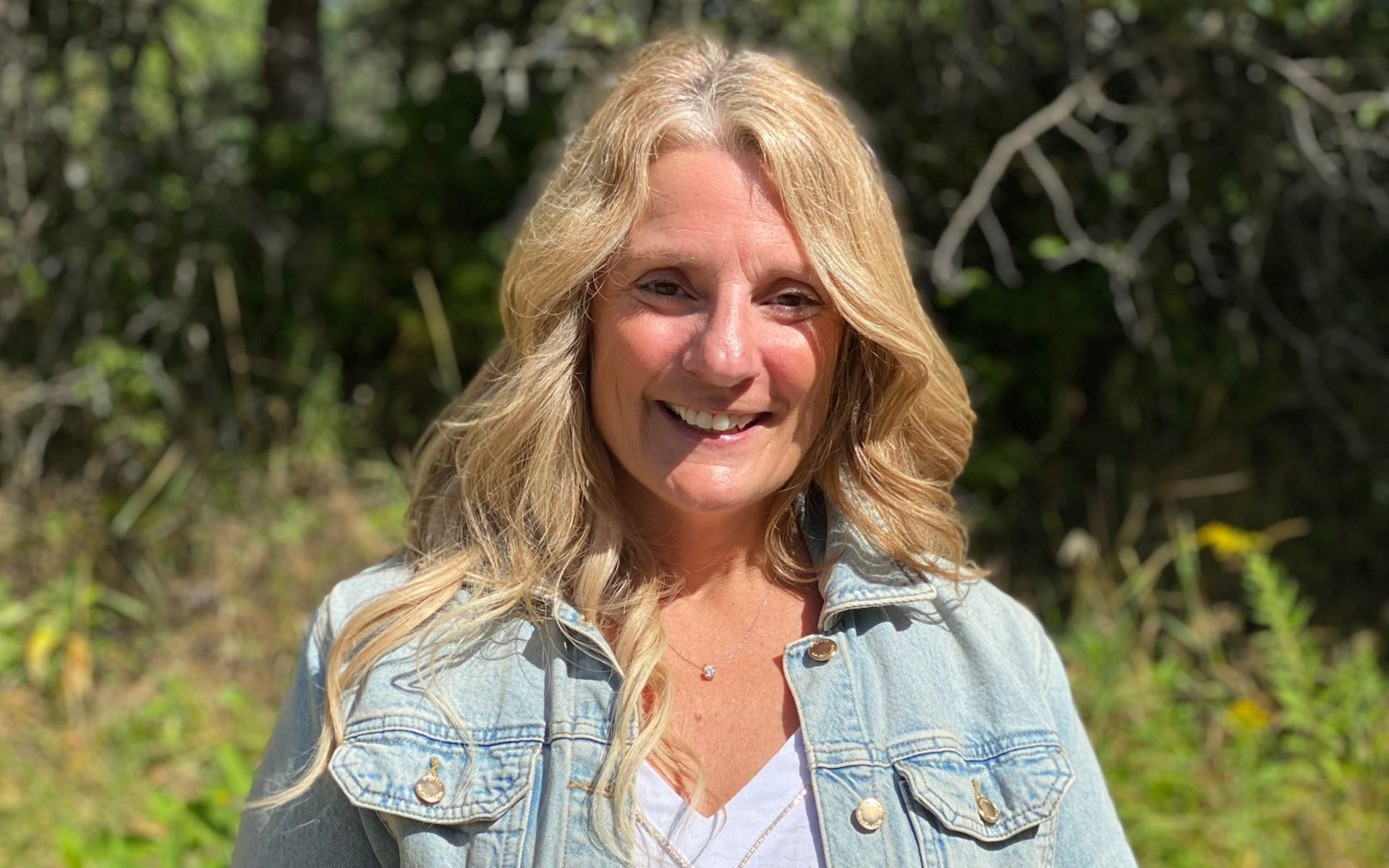 A photo of Darra; a woman with blonde hair smiling, wearing a denim jacket in a natural outdoor setting.
