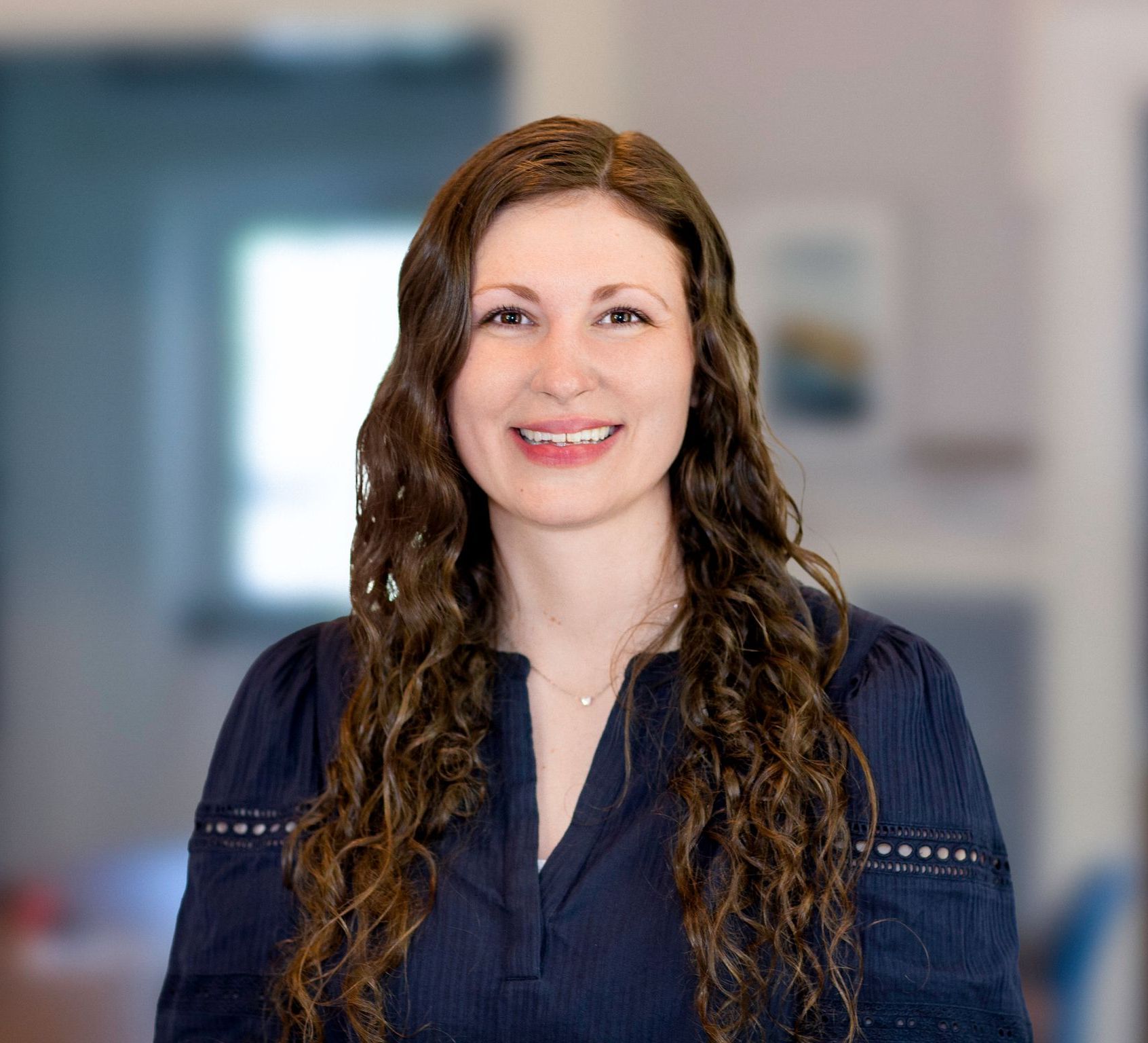 Woman with long, curly brown hair smiles at the camera, wearing a navy blue shirt, set indoors with a blurred background.