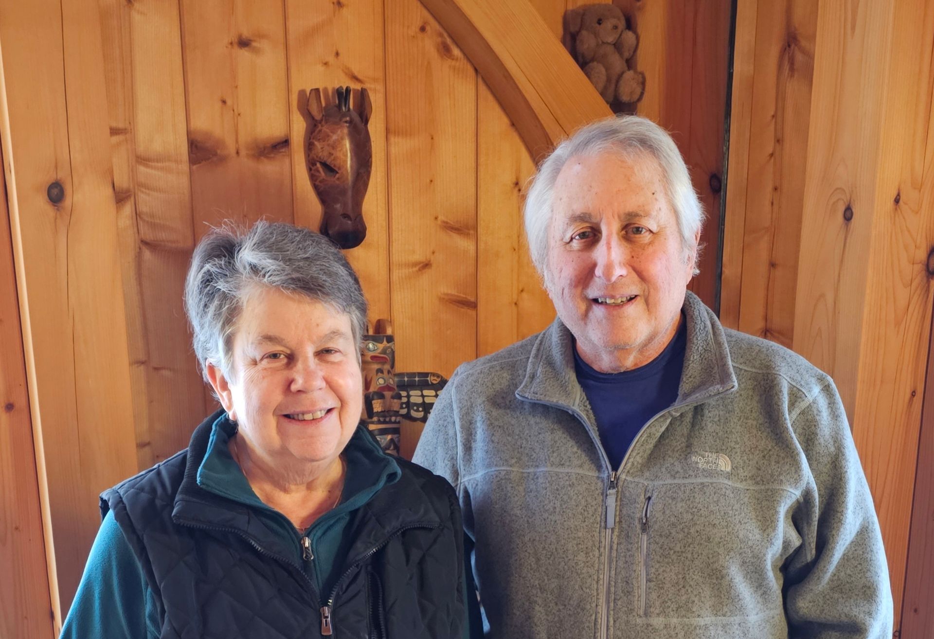 Two smiling older adults stand in front of a wood paneled wall. The woman wears a green vest, the man a grey zip-up sweater.