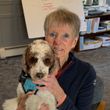 Woman smiles, holding a brown and white dog in a home office setting. The dog wears a blue harness.
